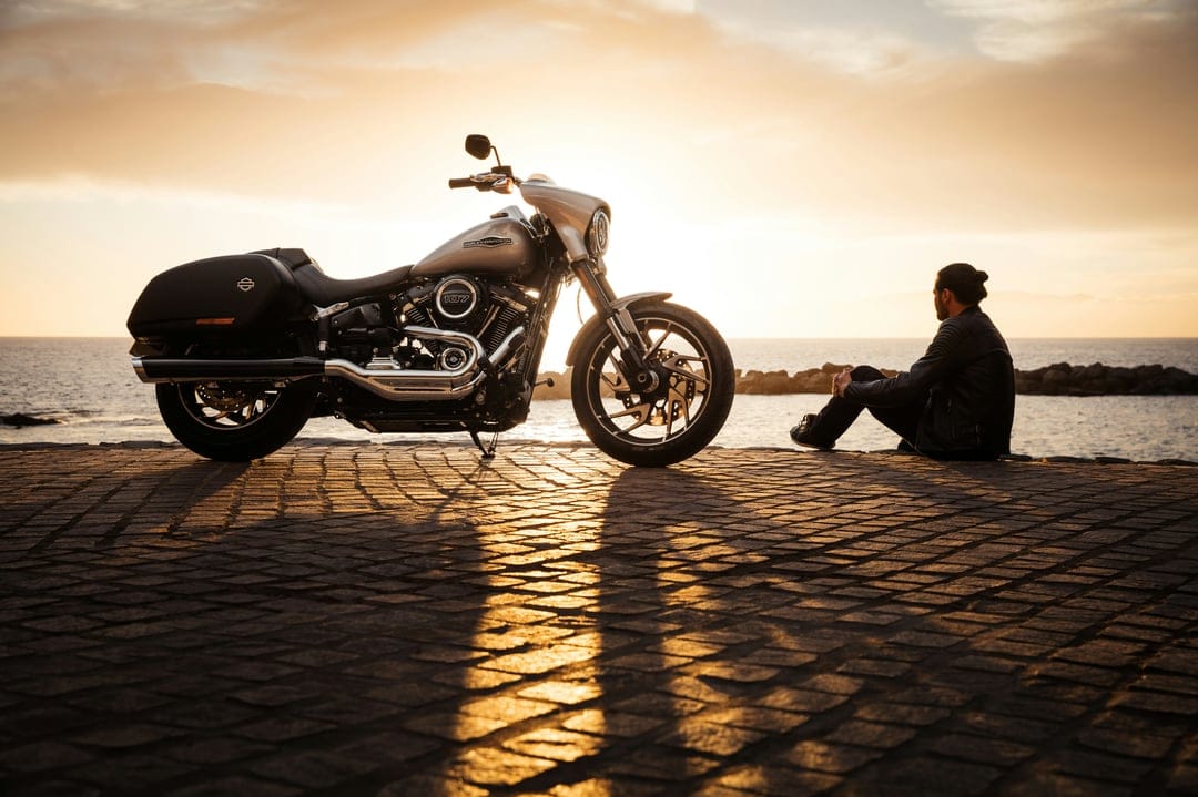 Man sitting on a stone path beside a motorcycle, watching the orange sun set over the distant ocean horizon.