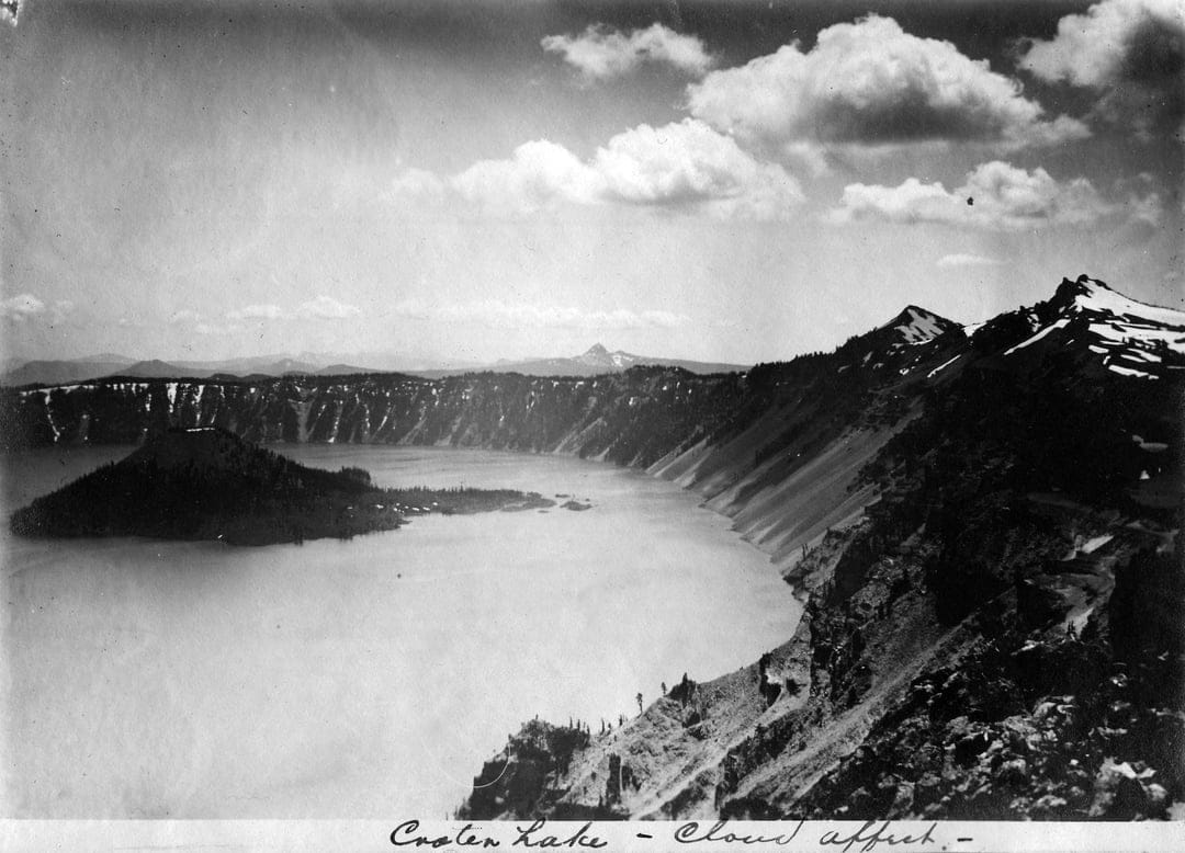 Wizard Island sits in the center of Crater Lake with steep caldera walls under a sky of dramatic clouds.