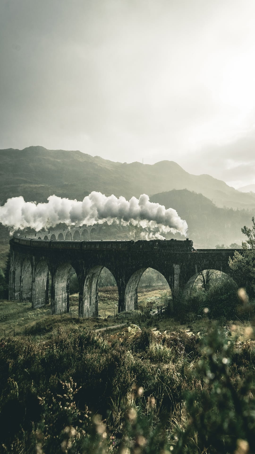 Steam locomotive billowing smoke while crossing a stone viaduct against foggy mountains and green hills.