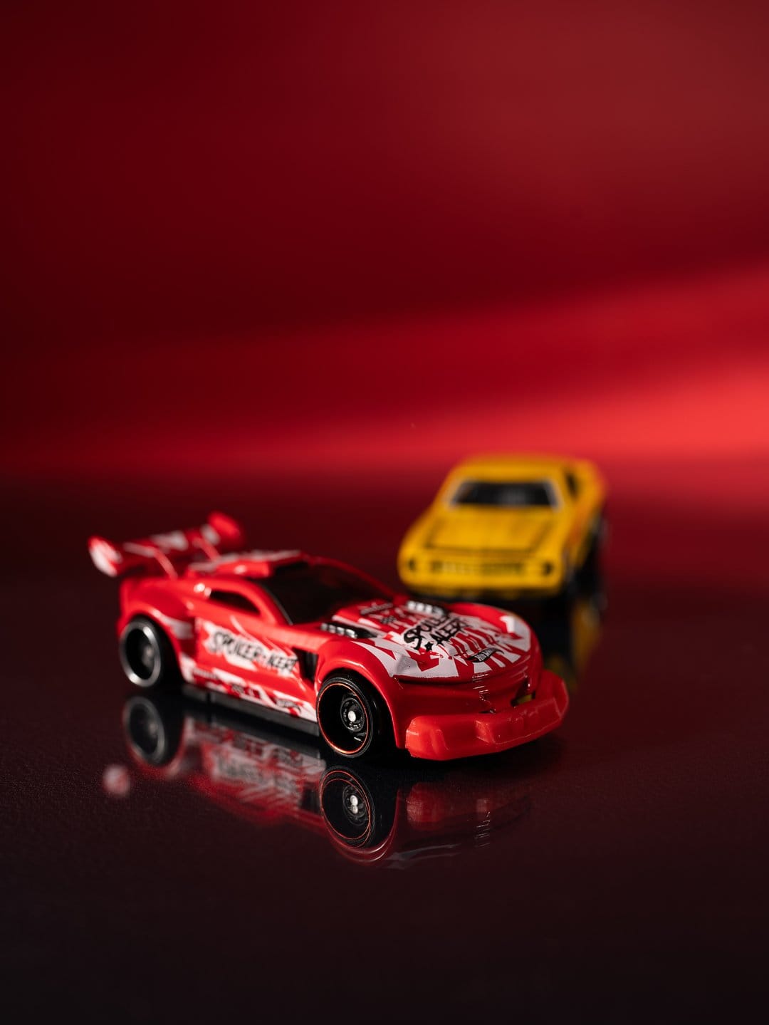 Red racing toy car with white stripes and spoiler on a reflective surface with a yellow car blurred behind it.