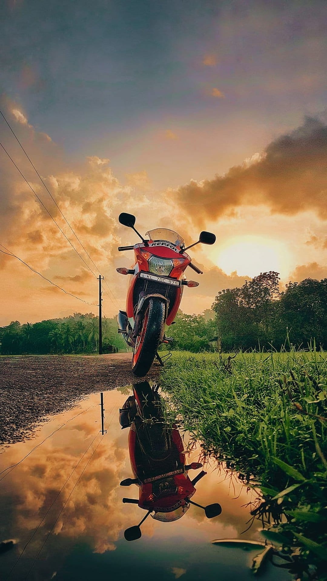 Red motorcycle reflected in a water puddle on a roadside during a dramatic orange and grey cloudy sunset.