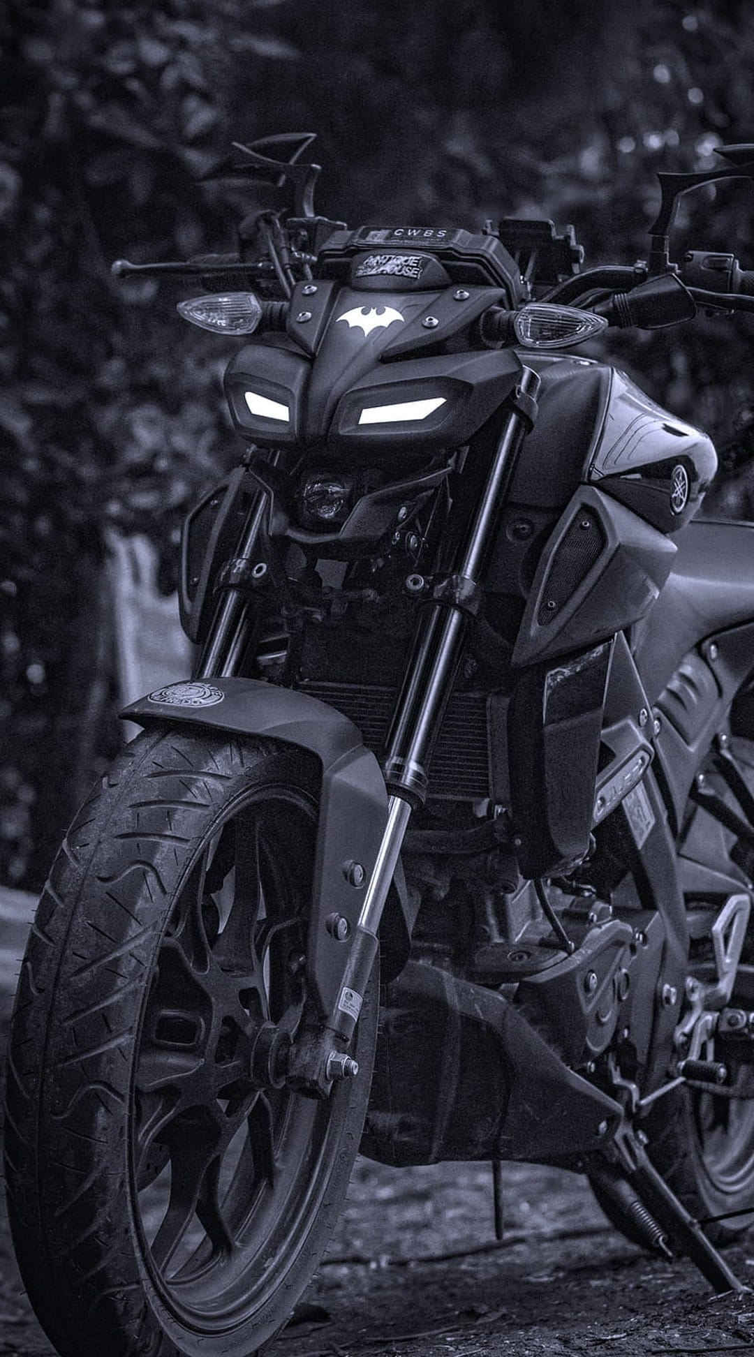 Matte black motorcycle front with a bat logo and glowing white LED headlights against a dark blurred background.