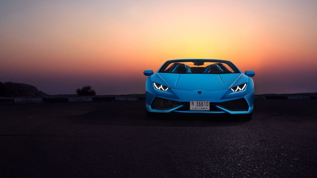 Blue Lamborghini Huracán Spyder convertible at sunset with glowing headlights on a dark asphalt road.