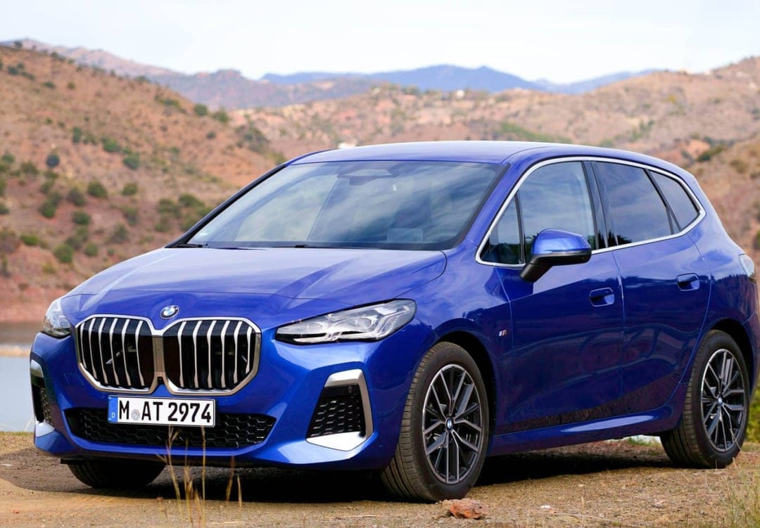 Blue BMW 2 Series Active Tourer parked on a dirt road with a backdrop of sunlit hills and mountains.