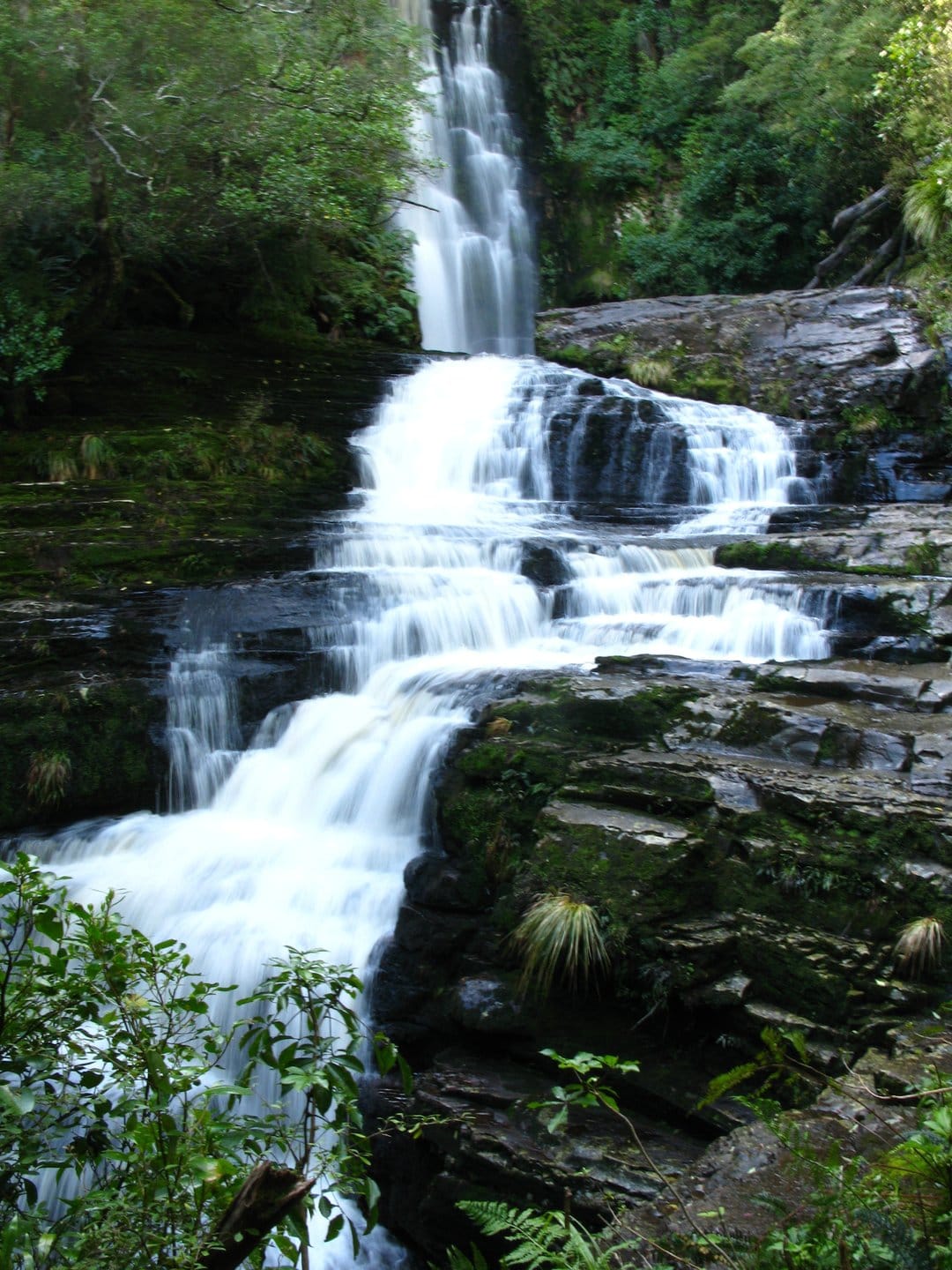 Tiered waterfall cascades over green mossy stones in a dense forest with a silky long exposure water stream.