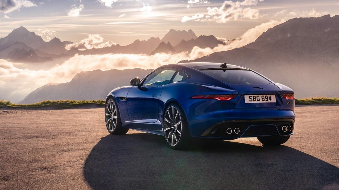 Blue sports car rear view on a mountain road at sunset with jagged peaks and white clouds in the distance.