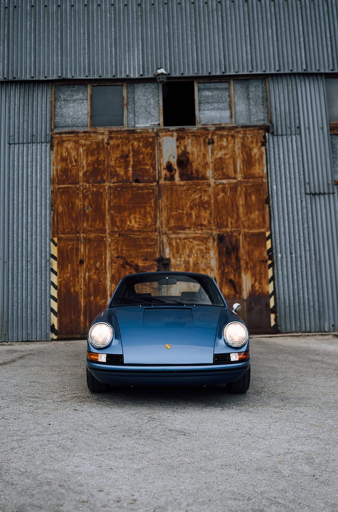 Blue Porsche 911 head-on in front of rusty corrugated metal doors on a weathered concrete industrial floor.