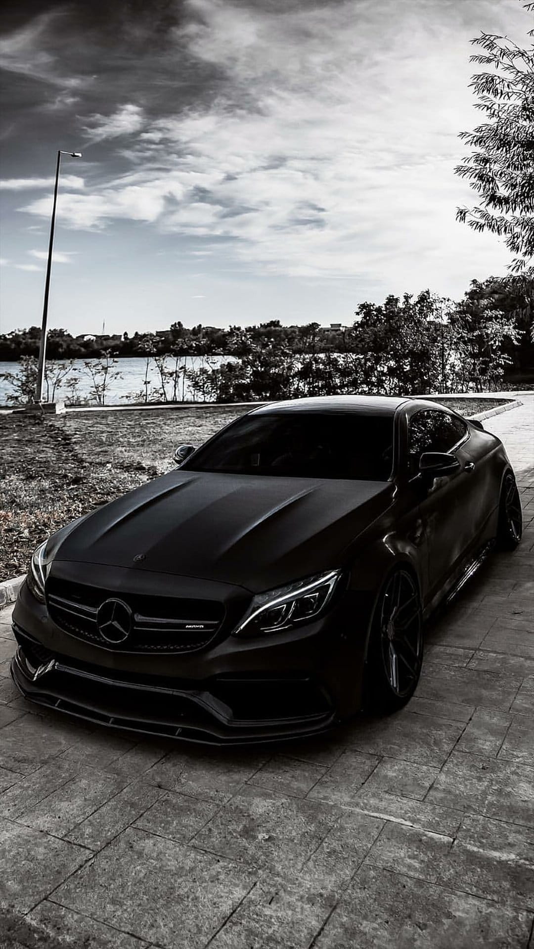 Matte black Mercedes-AMG coupe parked on a stone path by a tranquil lake under a bright blue sky with green trees.
