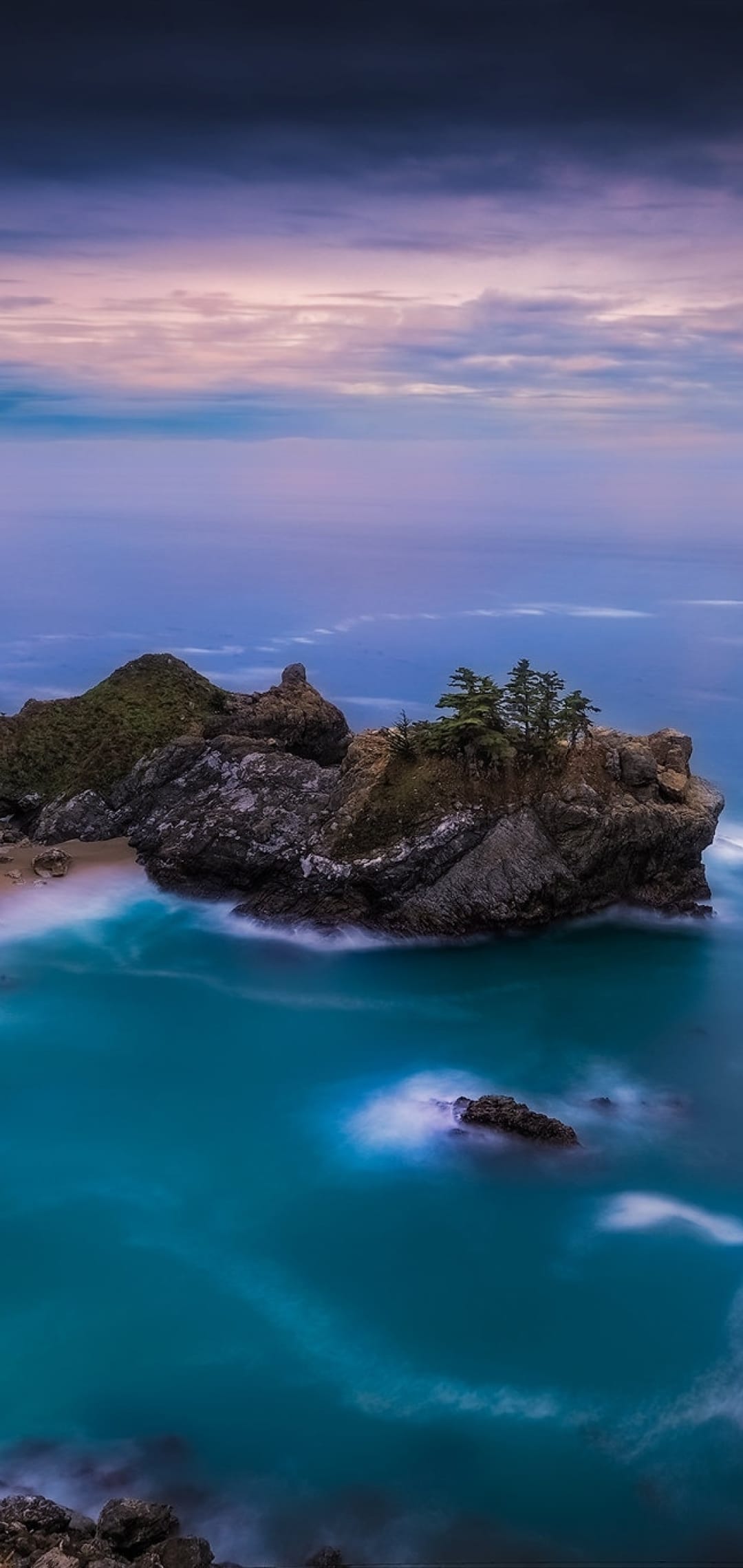 Emerald waters lap against rocky cliffs and a secluded beach under a purple and pink dawn sky in a vertical view.