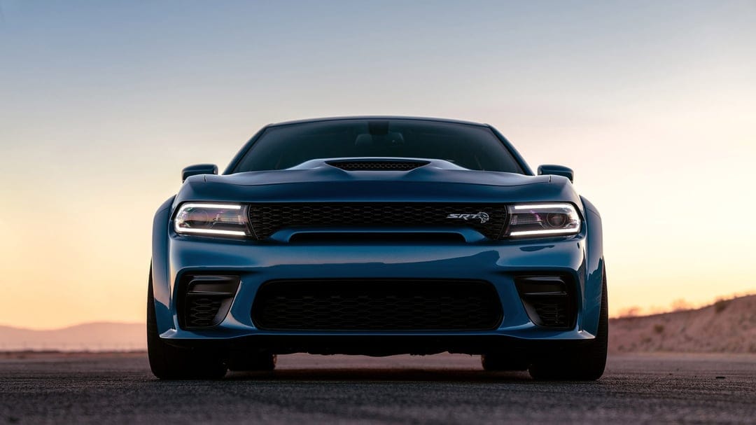 Front view of a blue Dodge Charger SRT Hellcat Widebody on a road at dusk with glowing LED headlights.