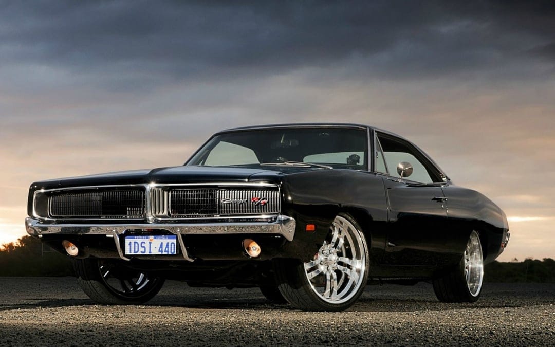 Black 1969 Dodge Charger R/T with chrome wheels on a gravel road under dark stormy clouds with sunset horizon light.