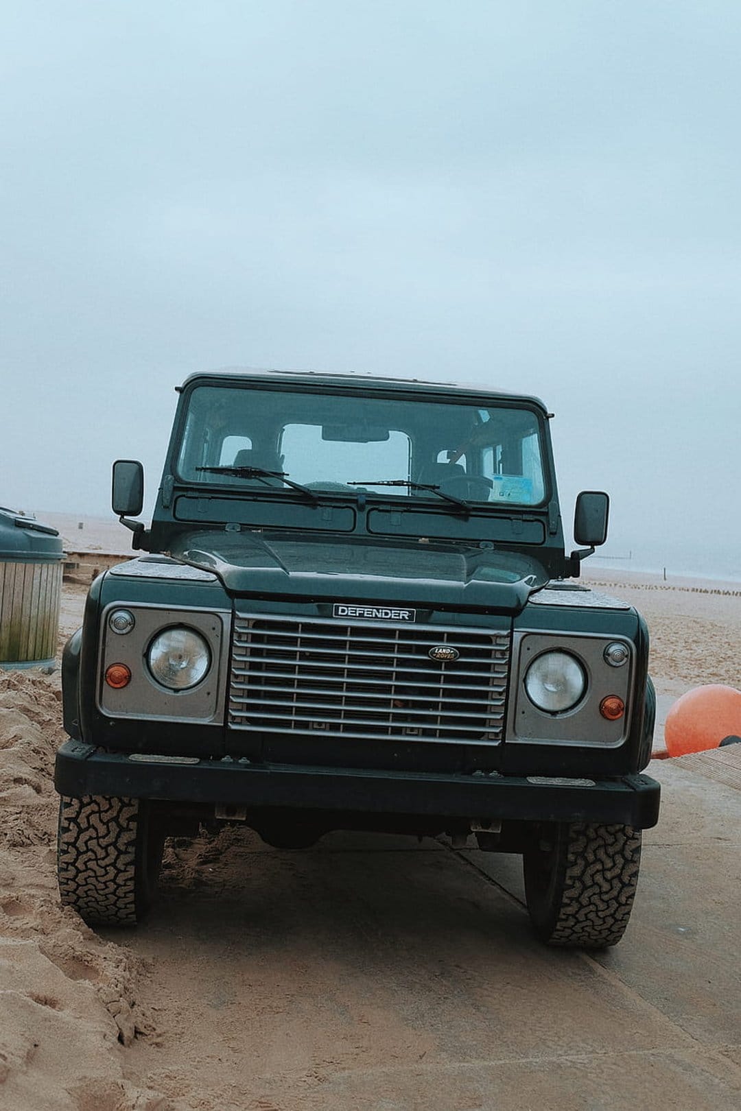 Dark green Land Rover Defender parked on a sandy beach under a moody overcast sky with rugged off-road tires.