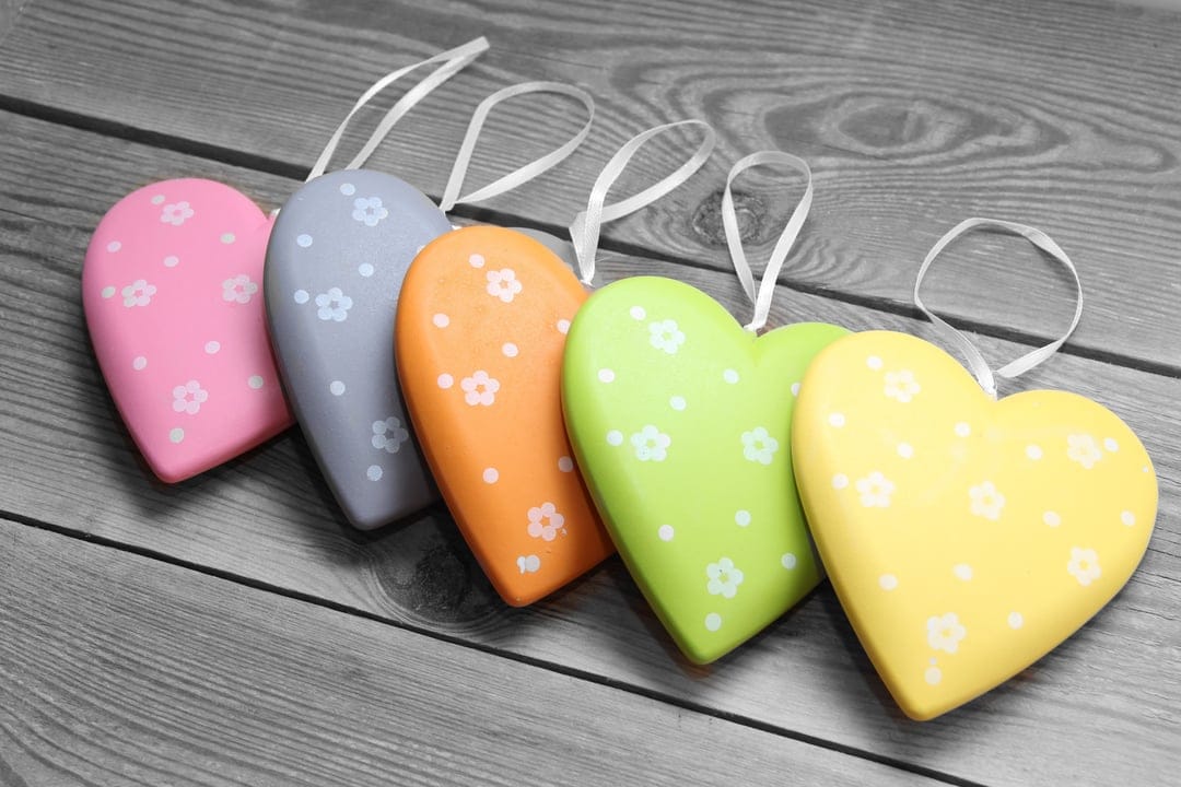 Colorful heart ornaments with floral patterns sit on weathered wood, tied with thin white hanging ribbons.