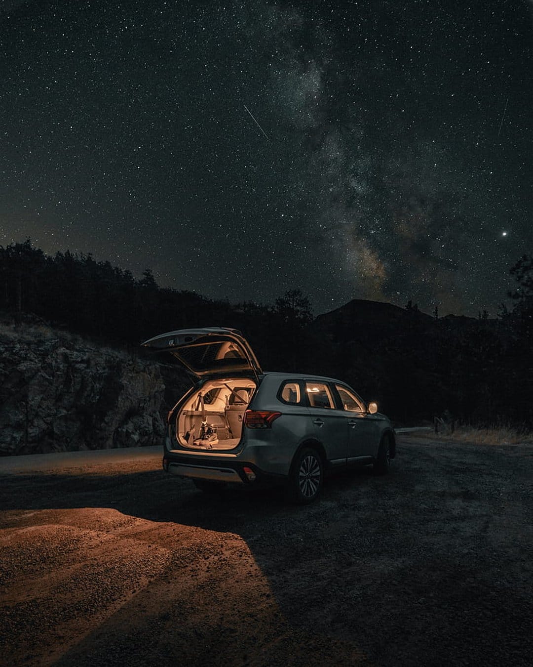 Silver SUV with an open tailgate parked on a dirt road under a starry sky featuring the Milky Way galaxy.