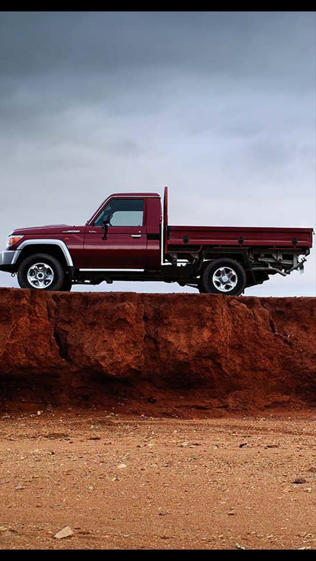 Red Toyota Land Cruiser pickup truck parked on a dusty cliff edge under a moody, overcast sky with rocky ground.