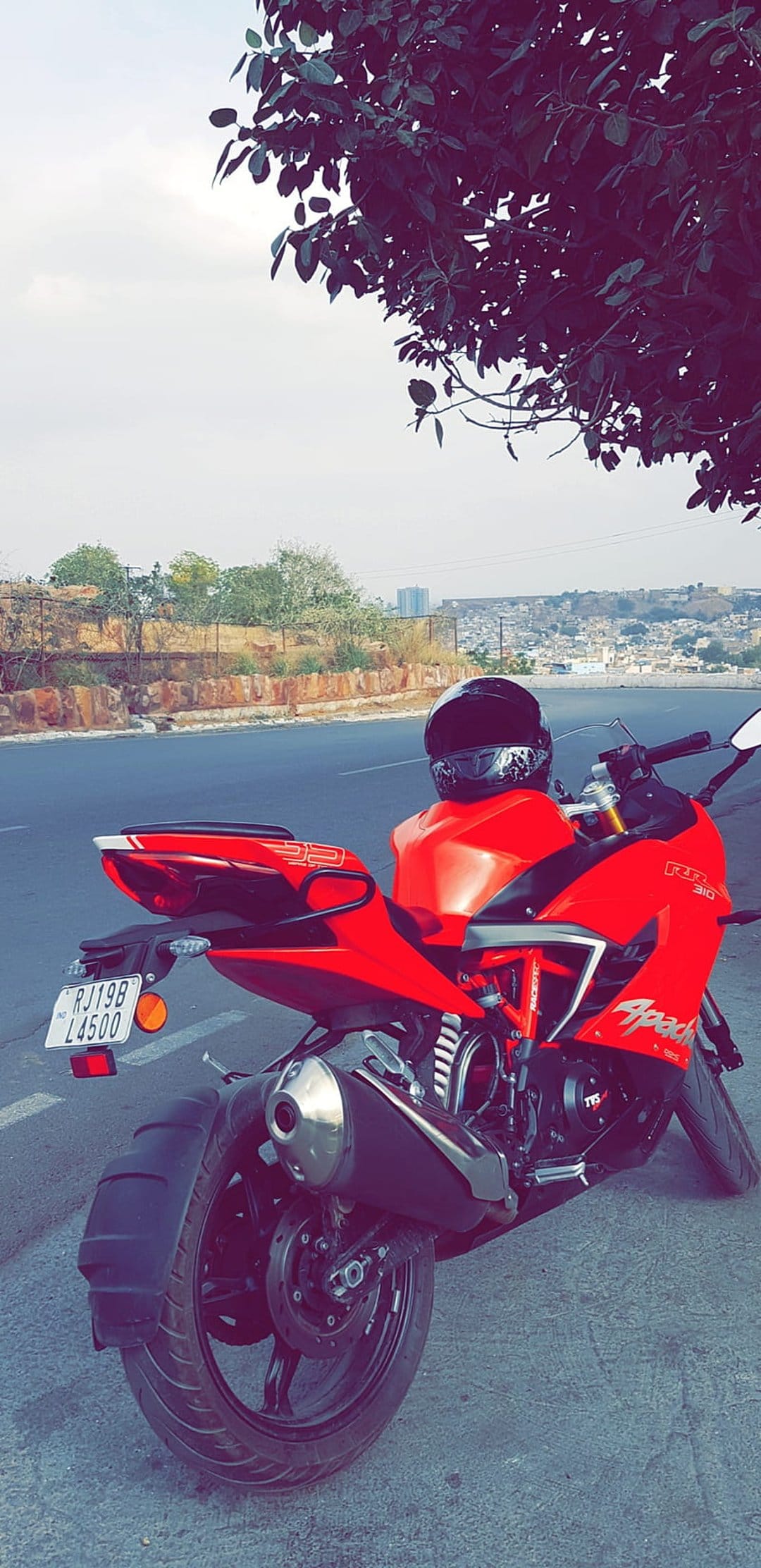 Red Apache RR 310 motorcycle parked on a roadside with a helmet on the seat, hills and trees in the distance.