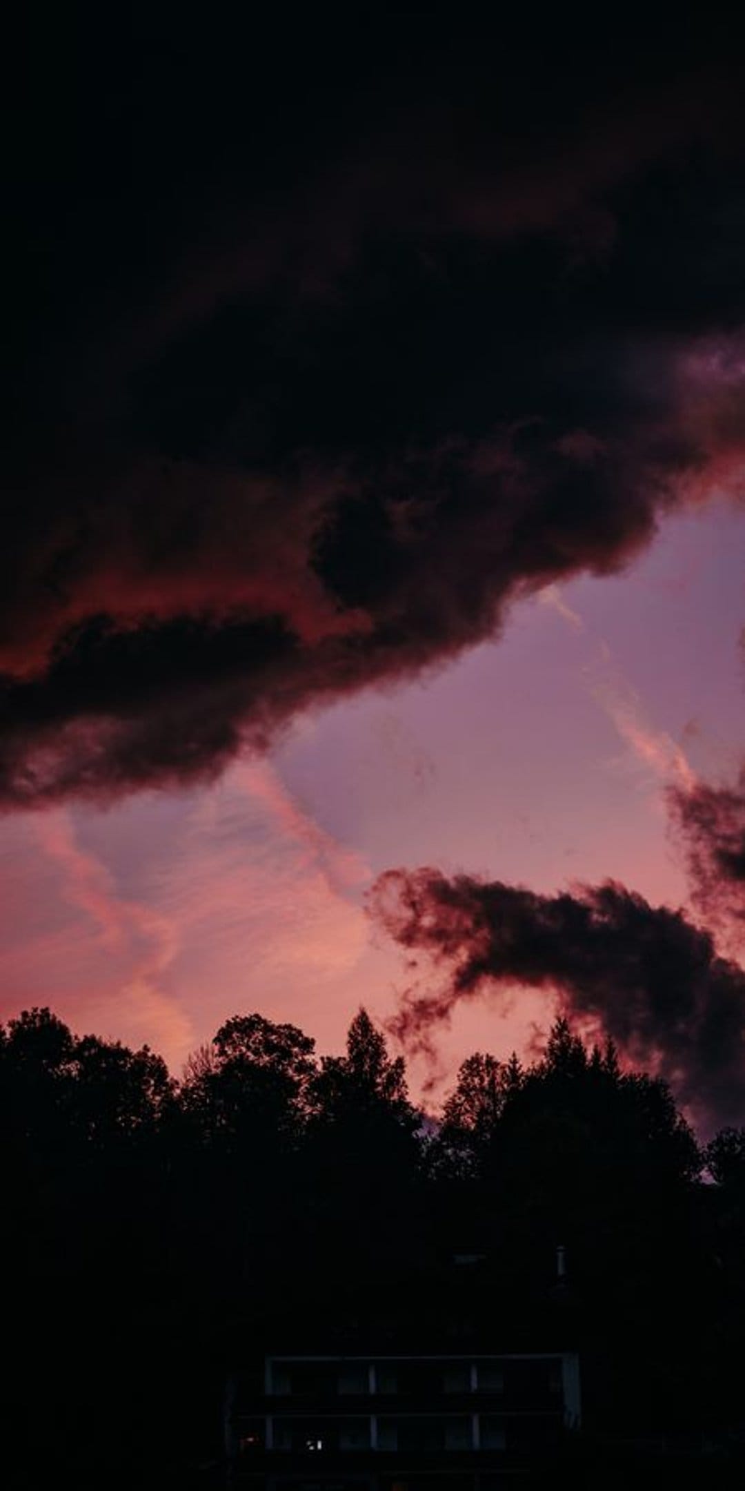 Deep purple and pink clouds swirl above a dark forest silhouette and a small building under a twilight sky.