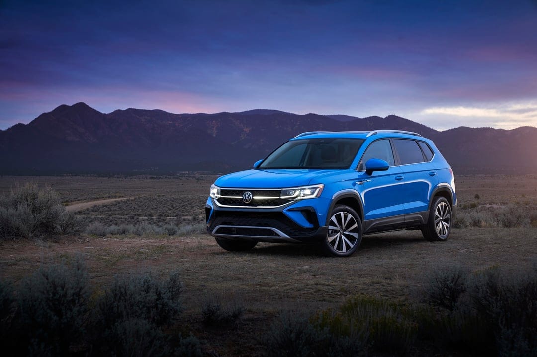 Blue Volkswagen SUV with glowing headlights parked in a desert landscape during a purple mountain sunset.