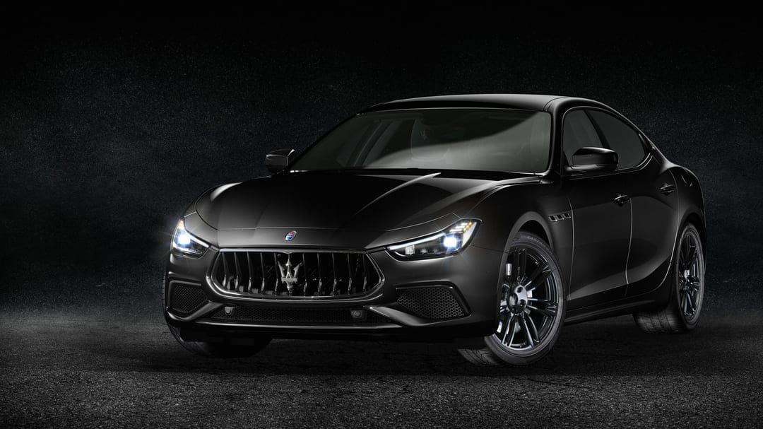 Black Maserati Ghibli sedan with glowing headlights on dark asphalt under dramatic studio lighting.