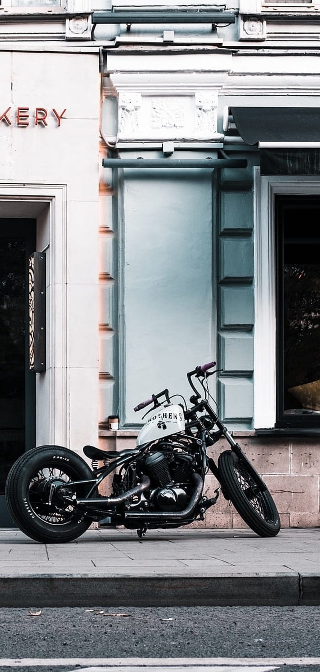 Custom black chopper motorcycle with white tank and purple handlebars parked on a sidewalk near a bakery sign.