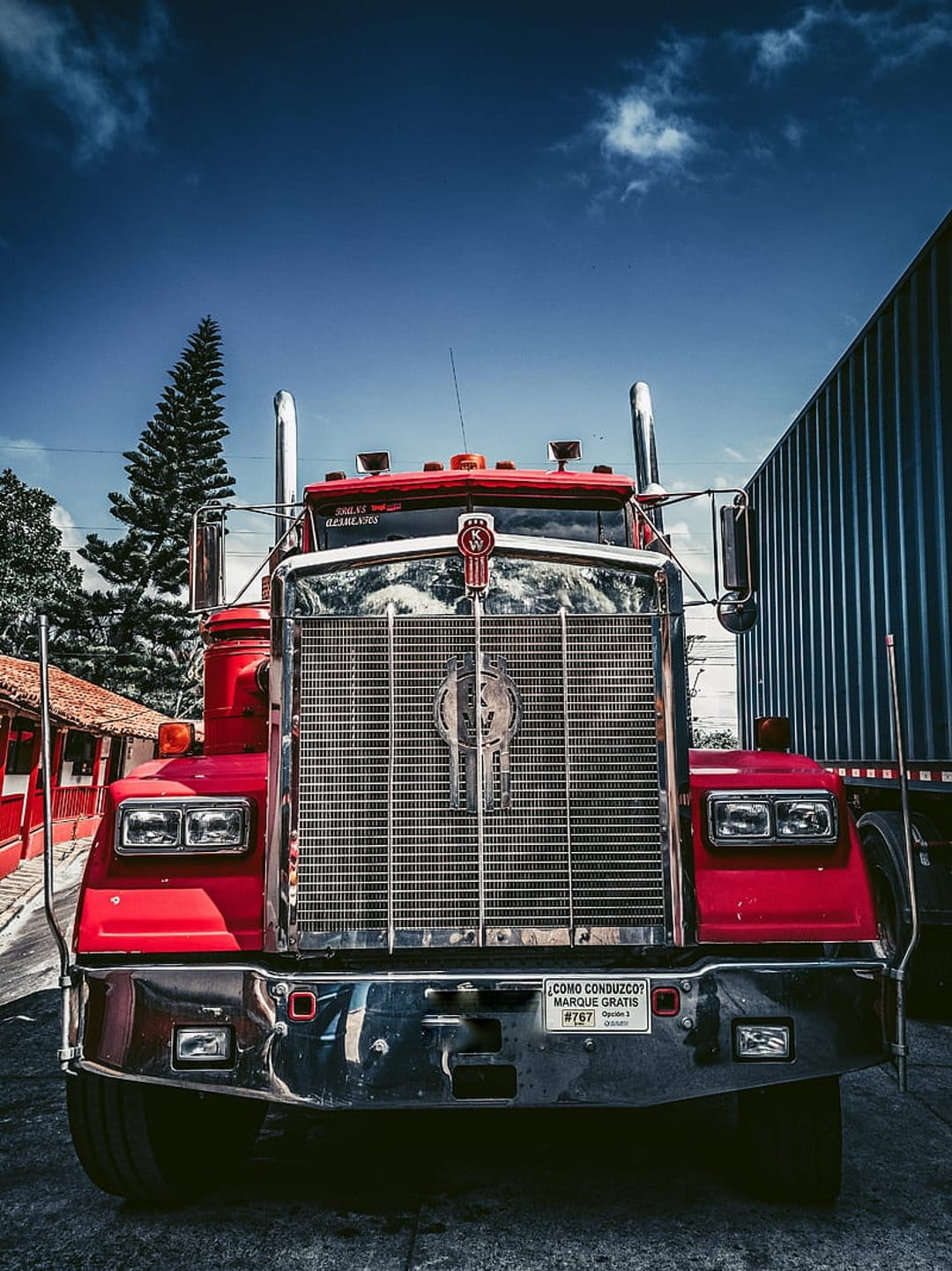 Red Kenworth truck front grille with polished chrome reflecting a blue sky and trees in a low-angle shot.