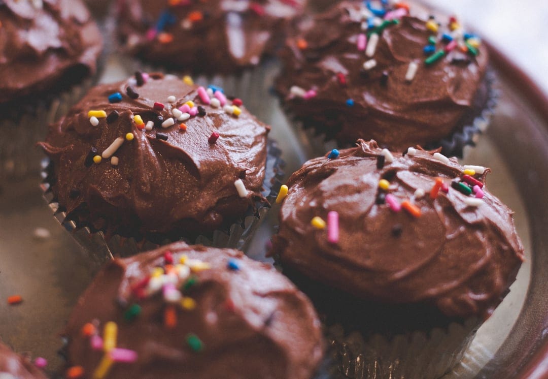 Chocolate cupcakes topped with dark cocoa frosting and rainbow sprinkles are arranged on a light-colored platter.