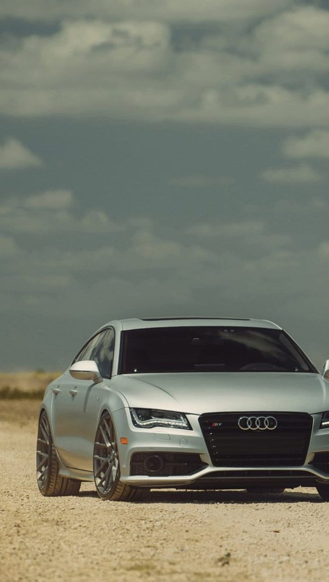 Silver Audi S7 parked on a gravel road featuring a prominent front grille and headlights against a cloudy sky.
