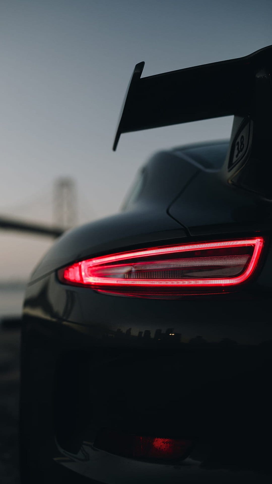 Porsche tail lights glow red on a black sports car with a blurred suspension bridge and city skyline at dusk.