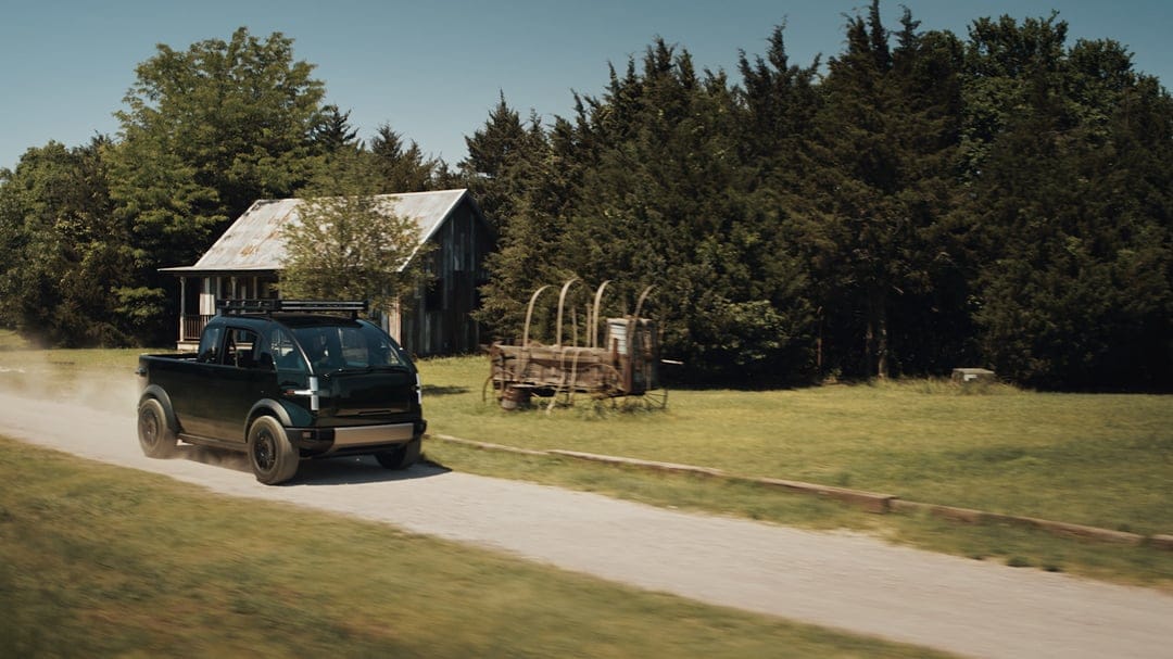 Dark green futuristic electric truck driving on a dusty rural road past a rustic wooden cabin and wagon.