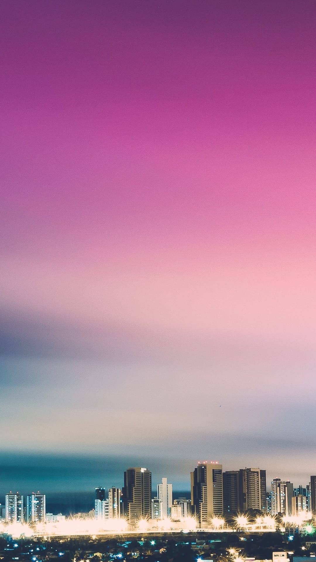 Shimmering coastal city buildings under a pink and purple sunset sky with glowing golden windows at dusk.