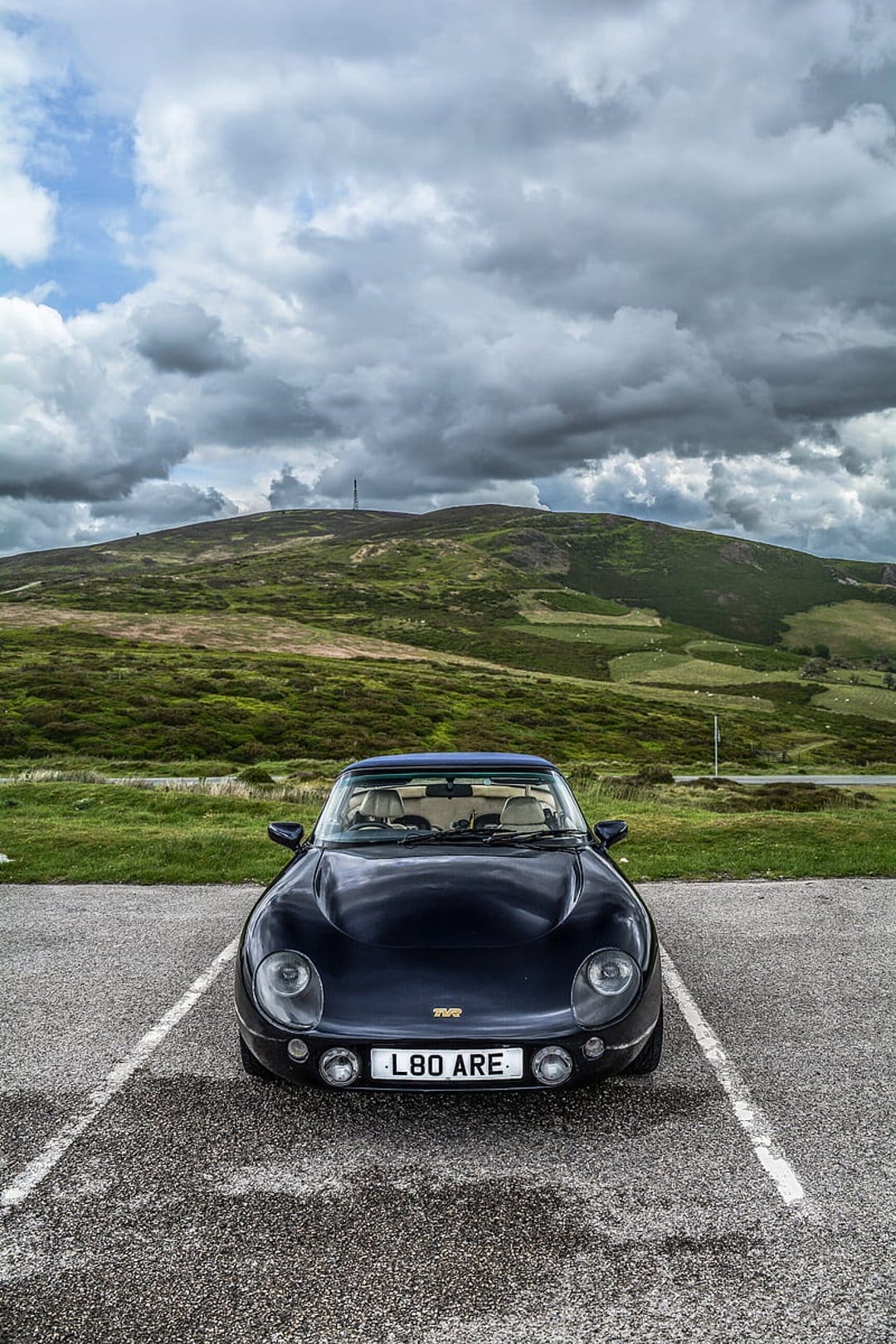 Dark TVR sports car parked on gravel with rolling green mountains and an overcast sky in the background.