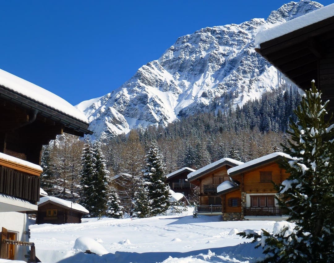 Timber chalets nestled in deep snow before jagged mountain peaks and evergreen pine trees under a clear blue sky.