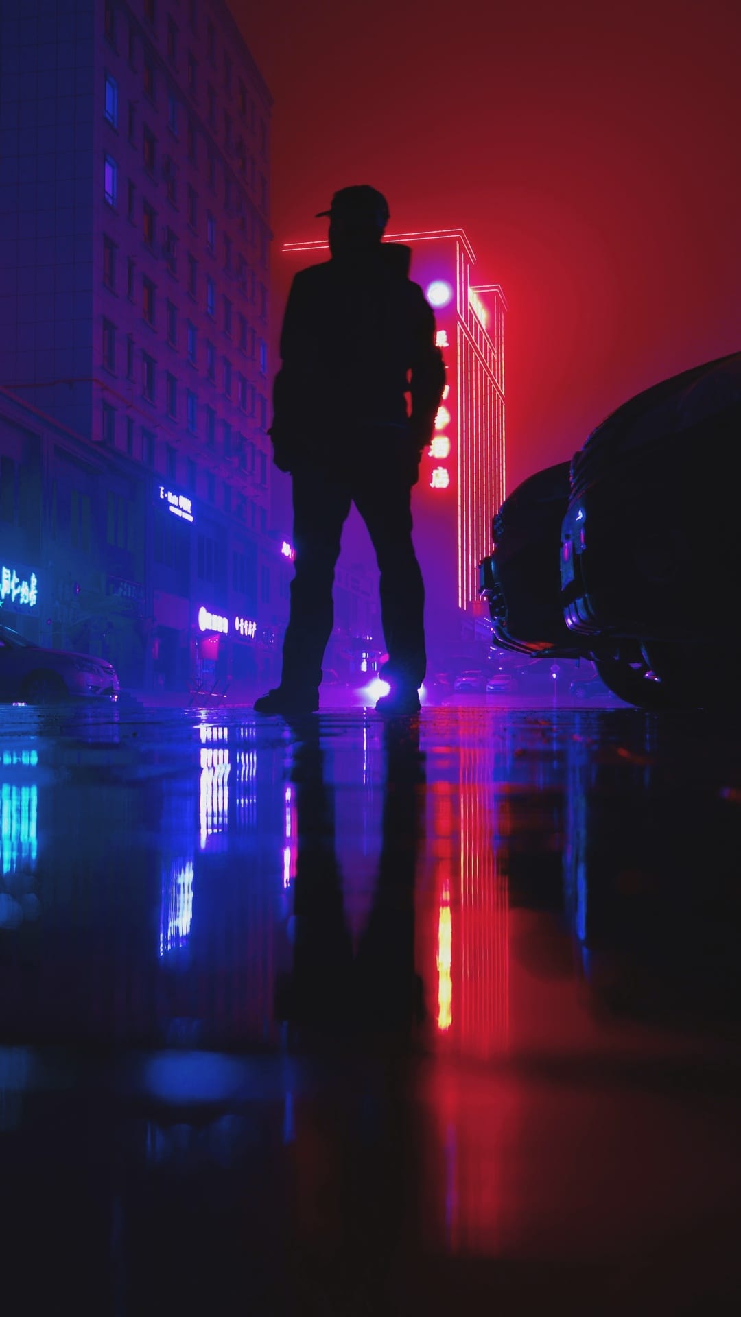 Capped person standing on a wet asphalt street reflecting vibrant red and blue neon lights from city buildings.