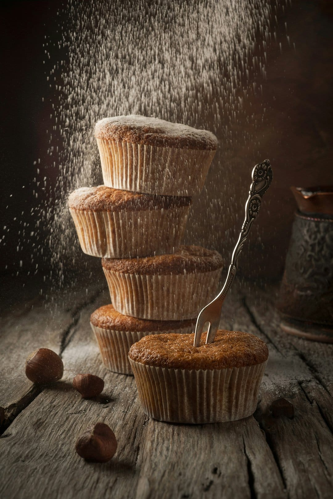 Muffins stacked on a wooden table are dusted with powdered sugar near a metal fork against a dark backdrop.