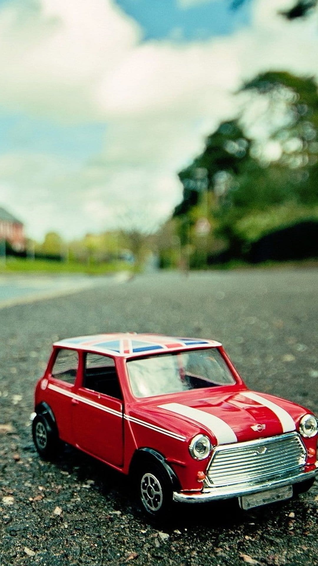 Red toy Mini Cooper car with white stripes and a Union Jack roof on a gravel road with a blurred background.