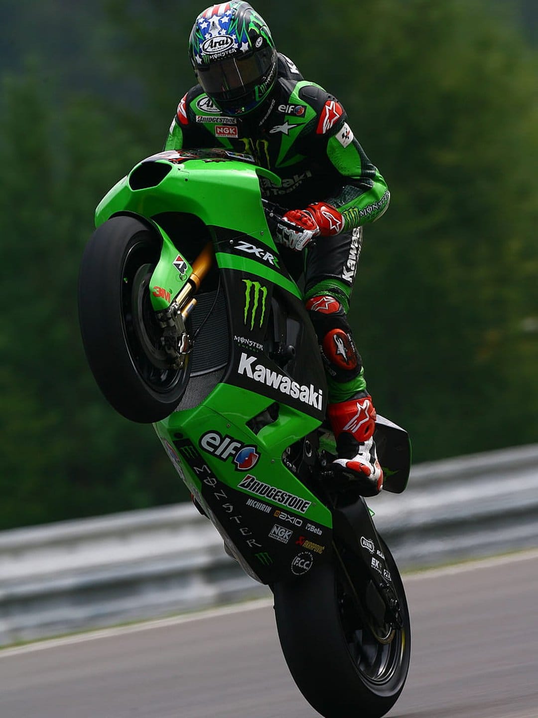 Motorcyclist on a green Kawasaki ZX-R performs a wheelie on a race track with an American flag helmet.