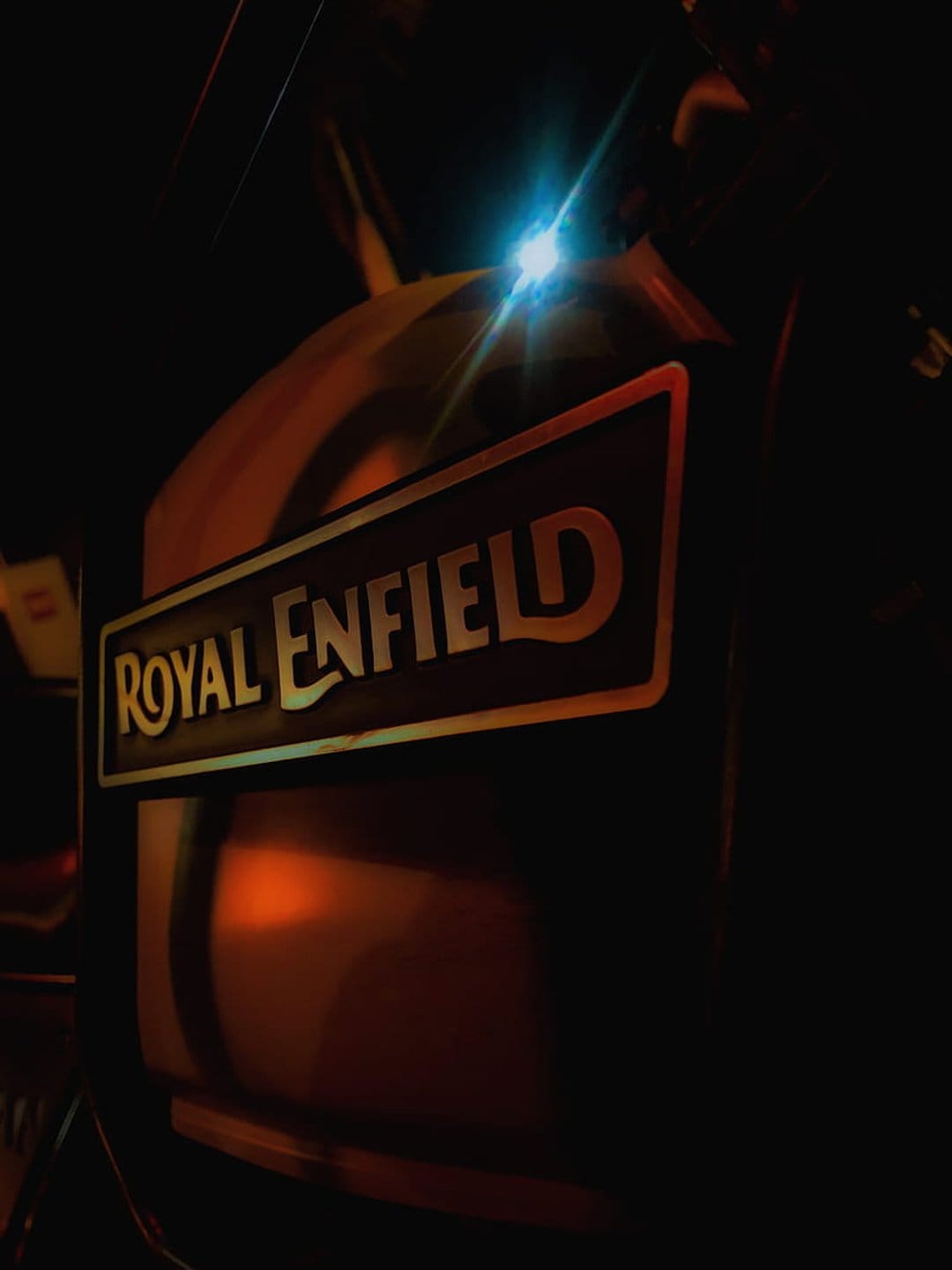 Royal Enfield chrome emblem glows under bright blue light with amber lens flares against a dark motorcycle frame.
