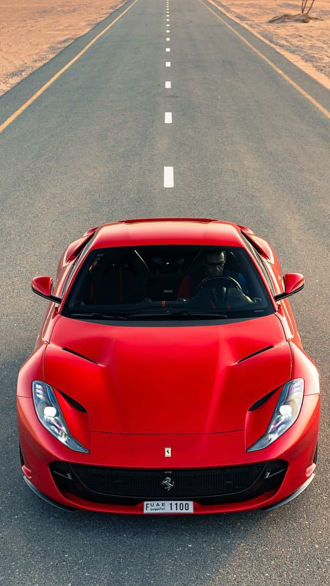 Red Ferrari sports car with a UAE license plate drives on a paved desert road surrounded by sand dunes.