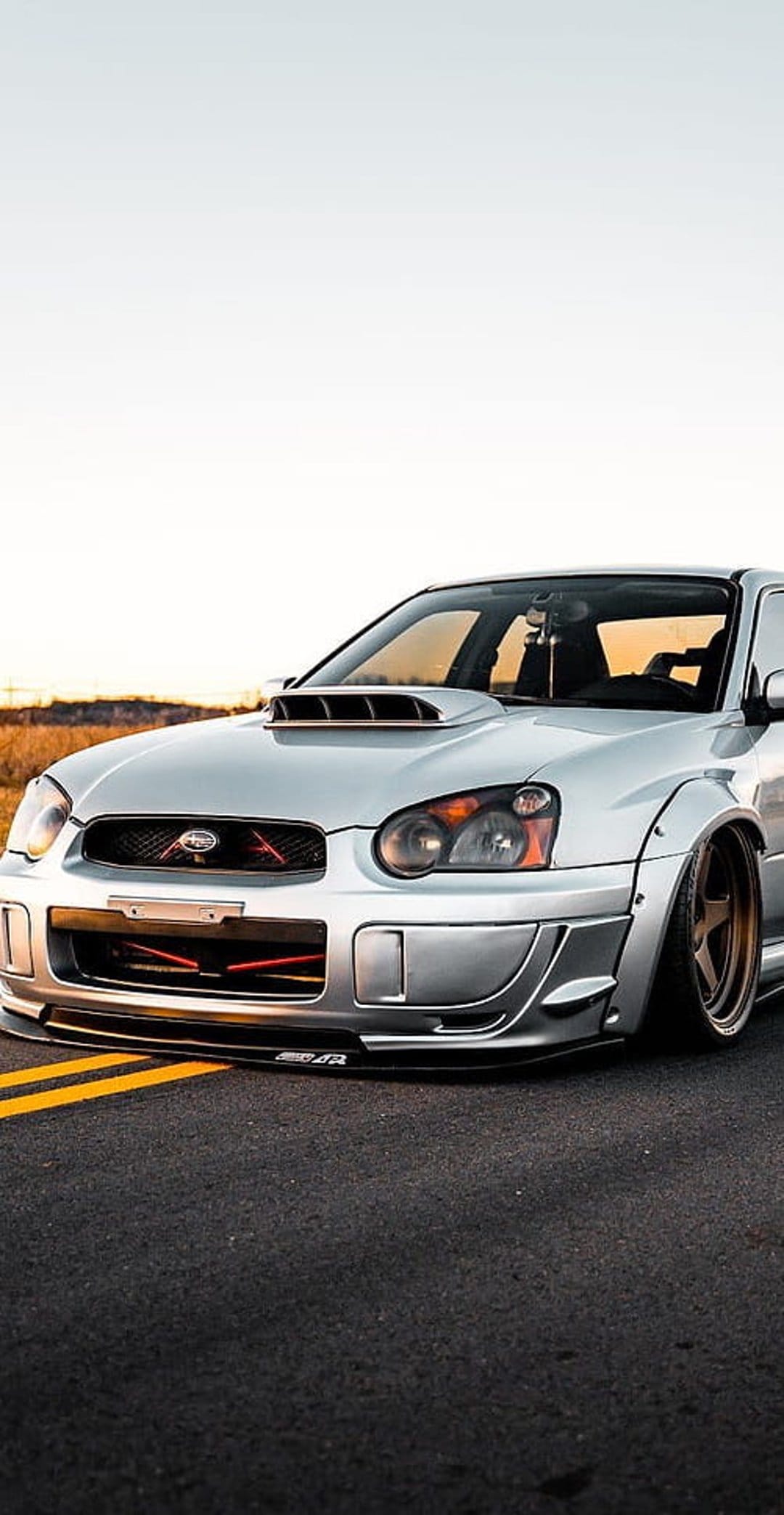 Silver Subaru WRX STI with a widebody kit and hood scoop parked on asphalt with yellow lane markings.