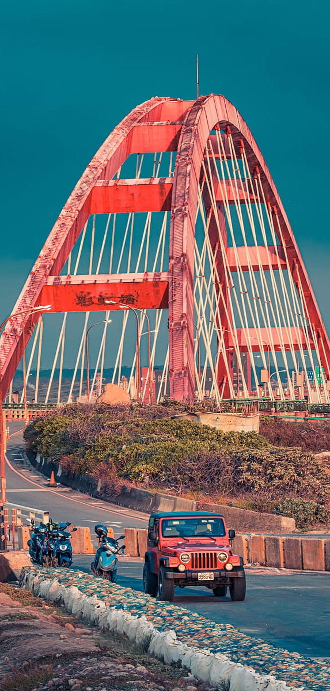 Red arch bridge with white cables towers over a coastal road featuring a red Jeep and green shrubbery.