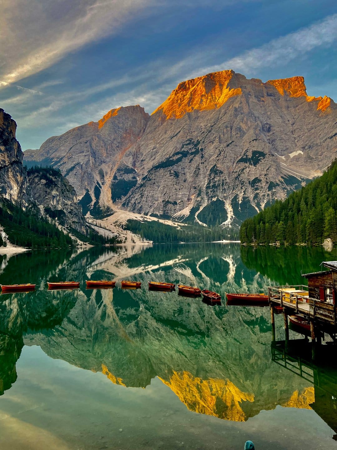 Wooden rowing boats anchor on a glassy emerald lake reflecting jagged sunlit mountain peaks and pine forests.