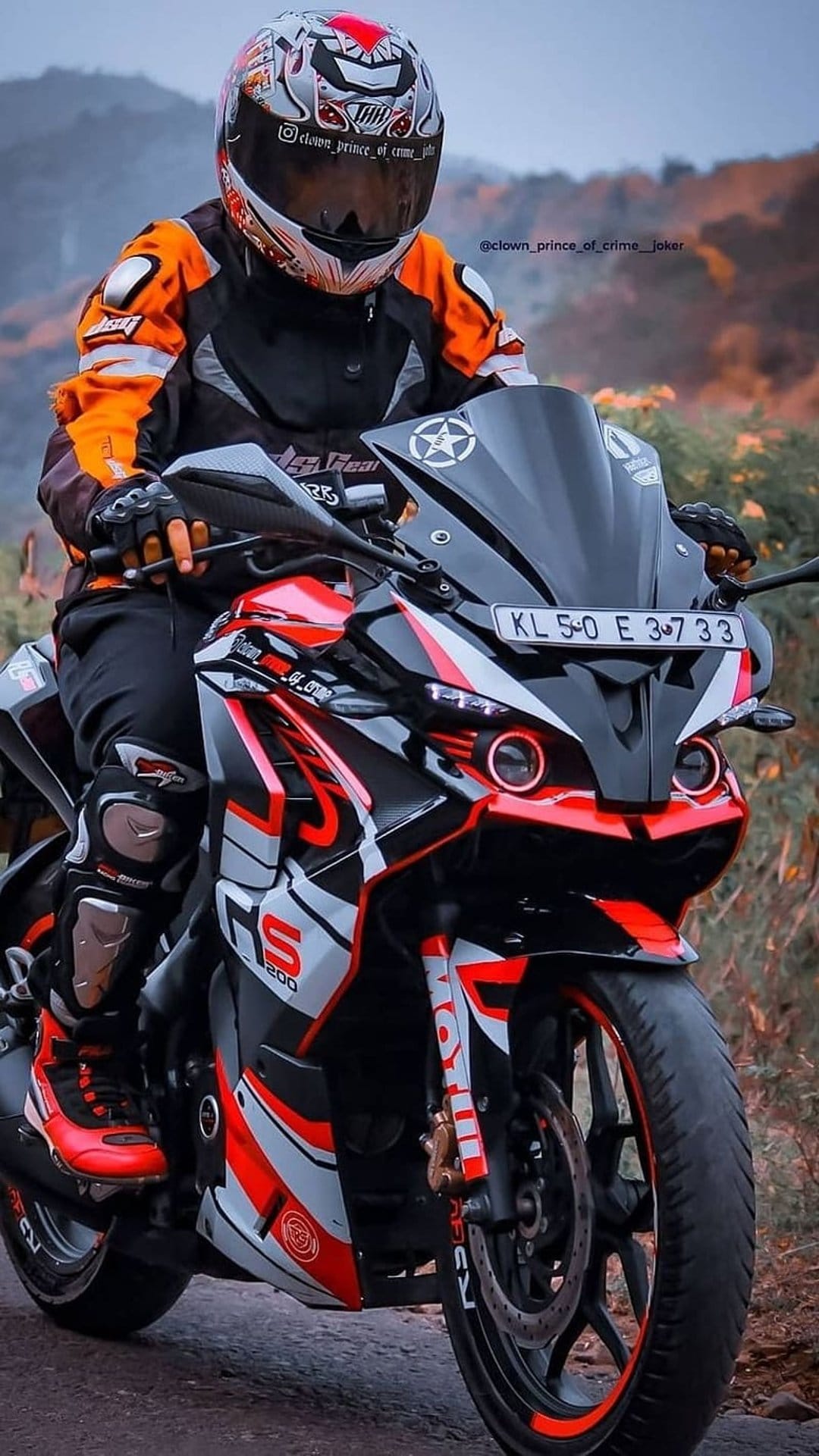 Motorcyclist in orange gear on a red sportbike sits against a blurred green forest backdrop on a country road.