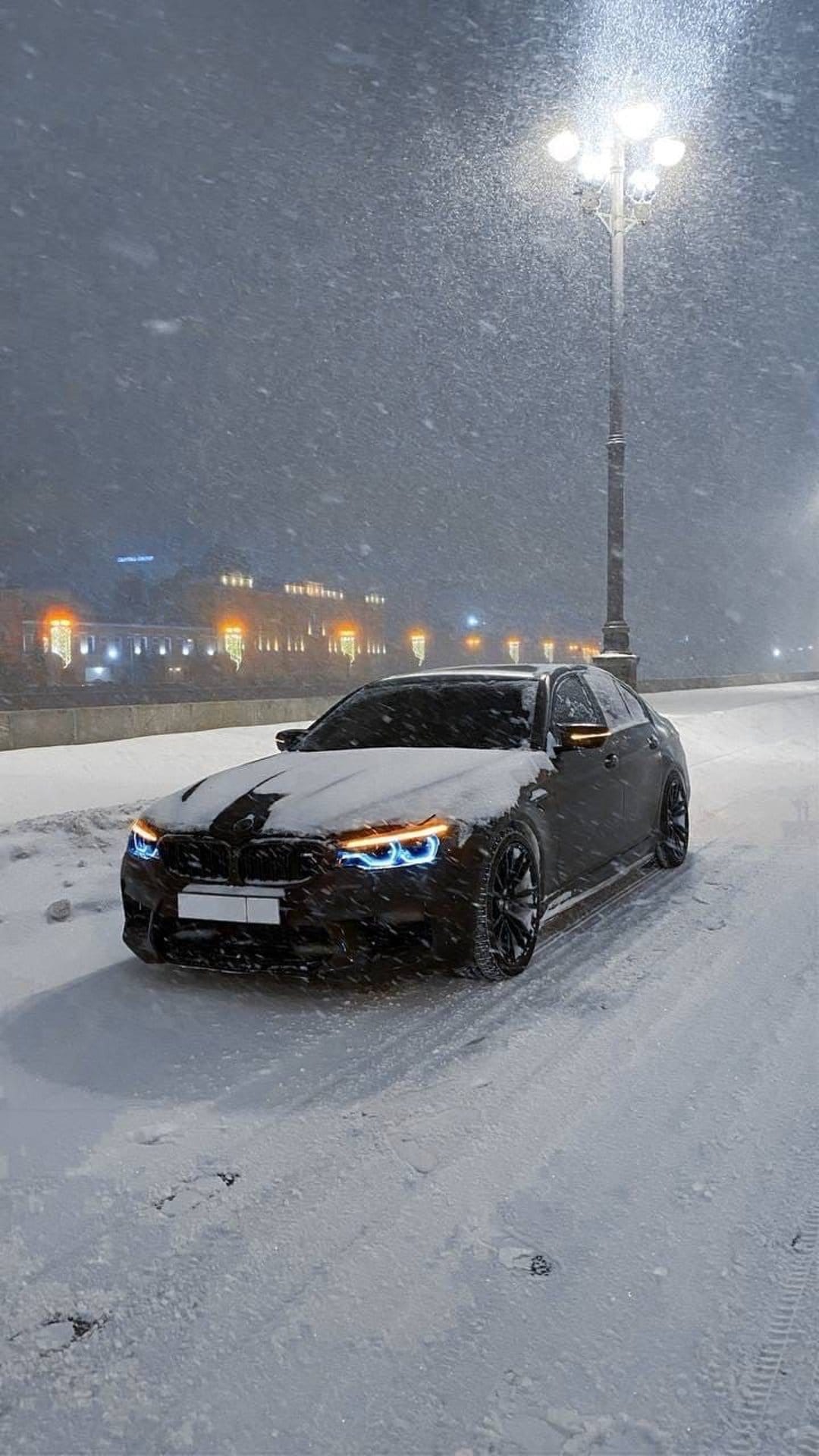Luxury dark sedan parked on a snowy road at night with glowing headlights and falling snow under streetlights.