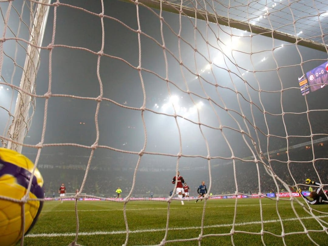 Soccer ball sits in a goal net on a misty green field during a night match under bright stadium lights.