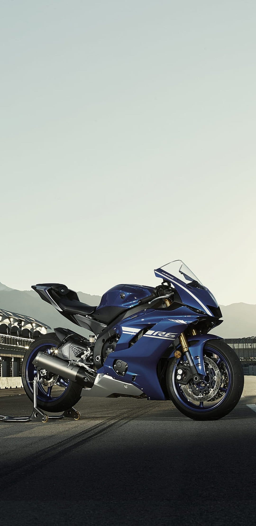 Blue Yamaha R6 sportbike on a paved racetrack with rugged mountains and a bright sky in the background.