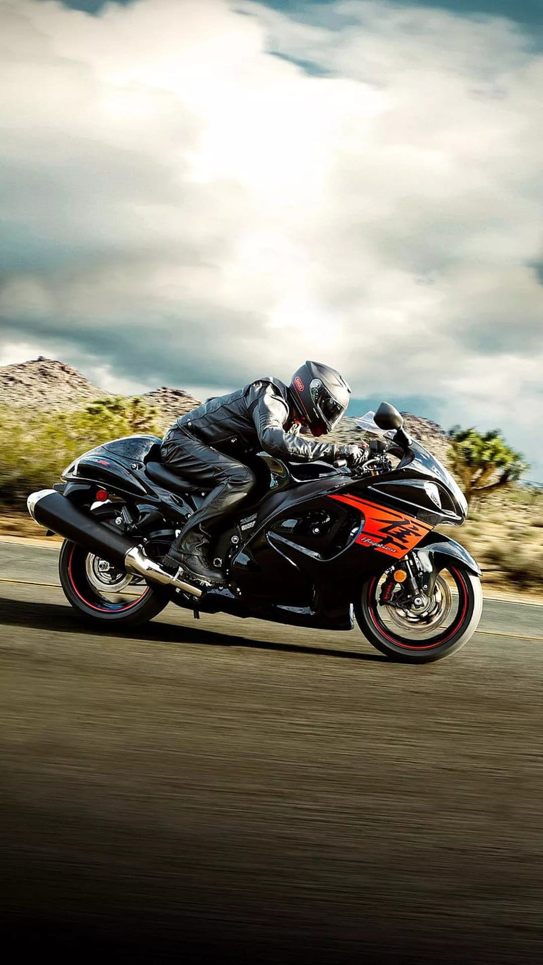 Motorcyclist on a black and orange sportbike speeds past desert mountains under a cloudy sky on a paved road.