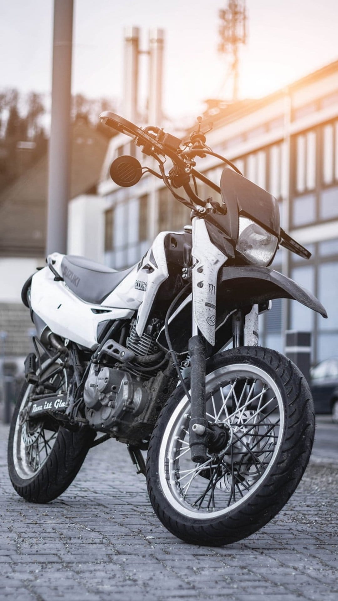 Suzuki dual-sport motorcycle with spoked wheels parked on a sunlit cobblestone street in an urban setting.