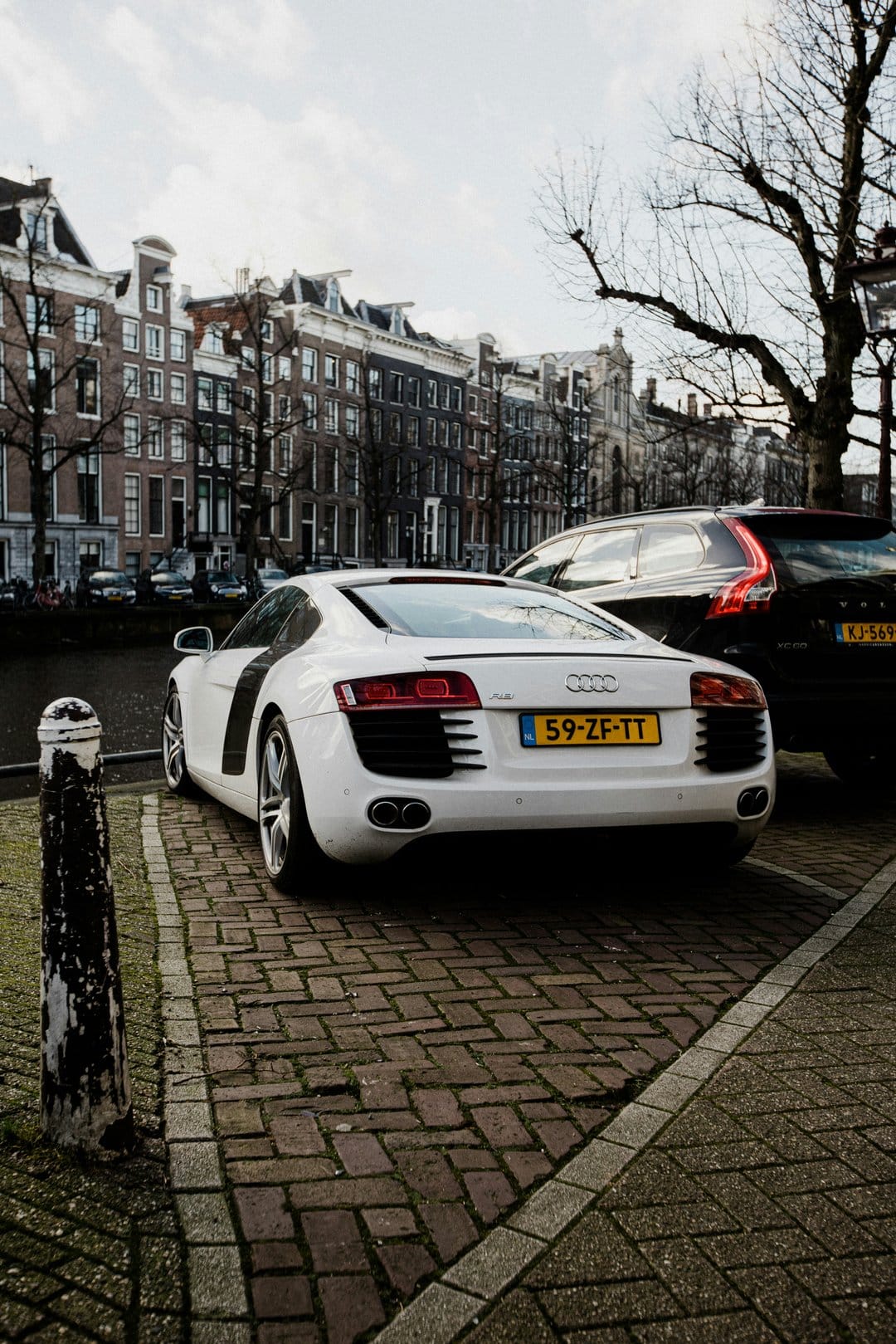 White Audi R8 sports car parked on a grey cobblestone street with historic Dutch canal houses in the background.