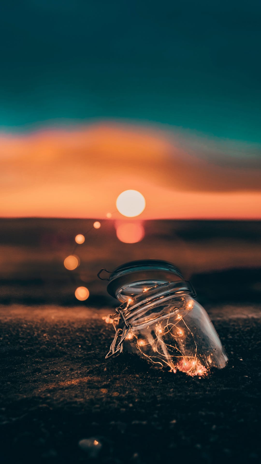Glass jar filled with warm fairy lights rests on dark ground before a glowing orange sunset and light trail.