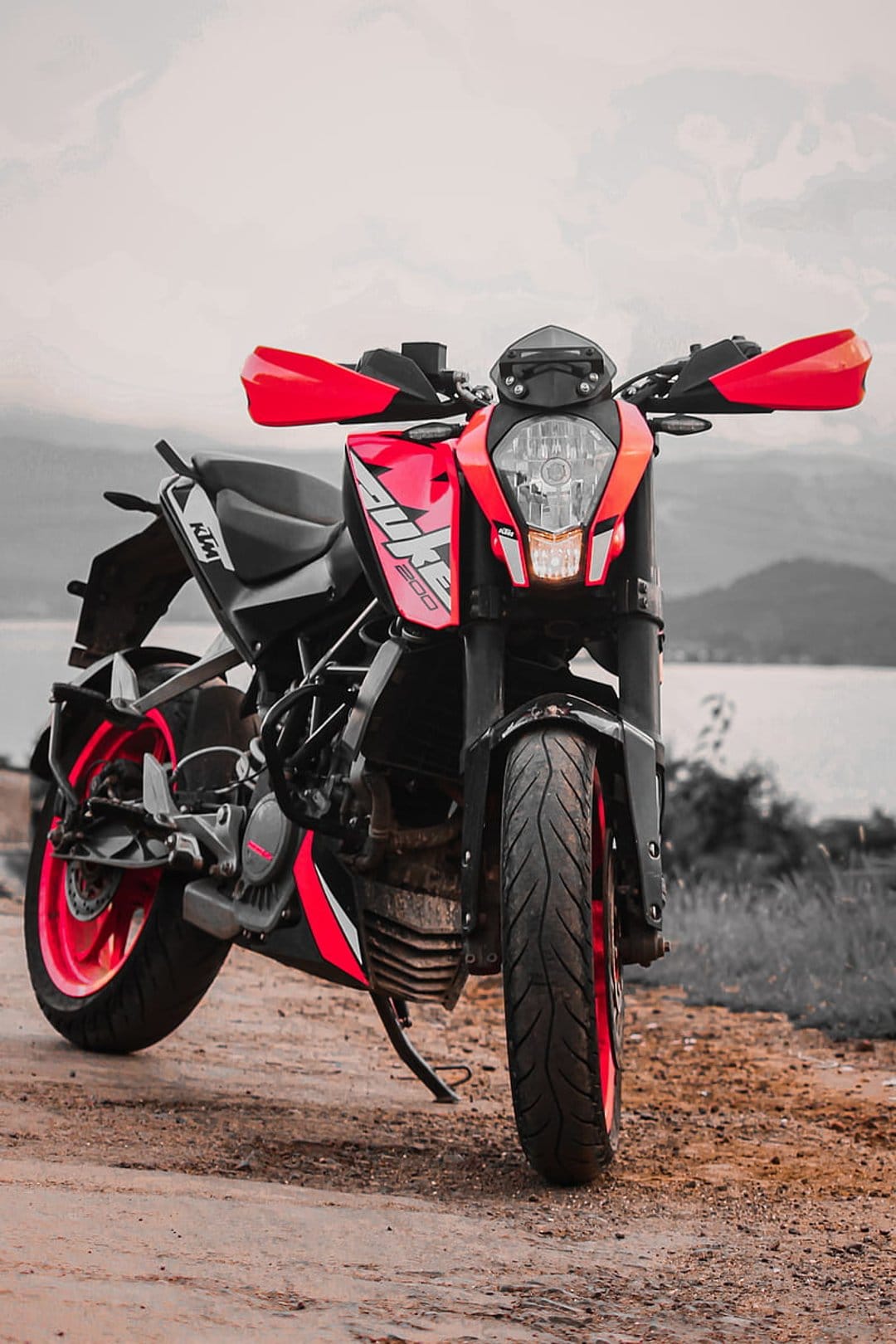 Red KTM Duke motorcycle parked on a dirt path with rolling hills and a calm lake under a cloudy sky background.
